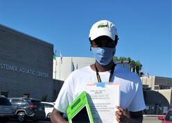 Gatete, wearing a cap and a mask holds health forms as he looks at the camera