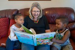 Julie Kurz reads to Sandra and Agape. They are sitting on a couch, the kids on either side of her with the book open on her lap.