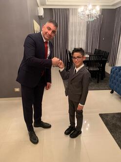 Maha's eldest son and 13-year-old grandson, both wearing suits and ties, fist bump and smile for the camera. 