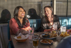 Two women laugh in a diner while sharing plates of food. 