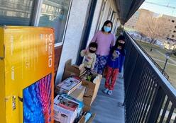 A mother and two children stand in front of the items they received through light one candle in front of their home