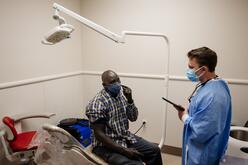 A dentist worker, holding a tablet is conversing with a man sitting on a chair in the dentist's office, who is pointing at his mouth