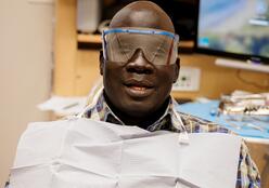 A man, wearing protective eye goggles and a paper bib, smiles at the camera from the dentist's chair