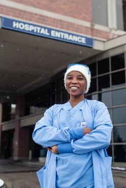 Nabila in front of the hospital where she works. She is wearing blue scrubs and a winter hat. 