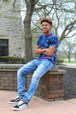 A young man leaning against an outdoor stone wall smiles with his arms crossed.