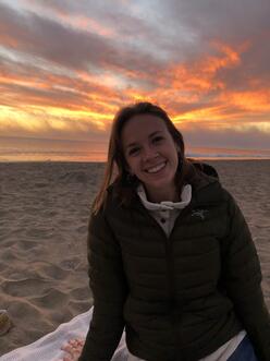 A woman in a coat sitting on a beach, with a colorful sunset in the background. 