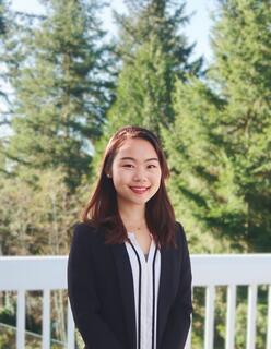 A woman smiles on a porch or deck, with evergreen trees in the background.