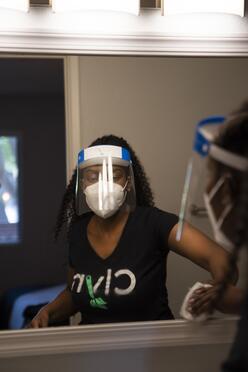 Diana cleans a window while wearing a shirt with the name of her app, Clyn, as well as a face shield and mask. 