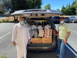 2 MCC members stand next to the back of a car with the hatch open to reveal food donations about to be delivered to families