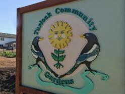 A wooden sign contains the words "Turlock Community Gardens" and has the image of the sun smiling while growing out of a plant. The sun is flanked by two magpies, which are common birds in the San Joaquin Valley.