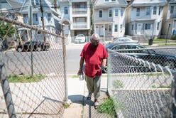 Congolese artist Muyambo Marcel Chishimba walks around a fence and down a paved path into his backyard in Elizabeth, New Jersey
