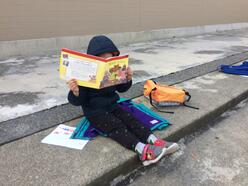 An elementary school student in a blue jacket sits on concrete steps, reading a book which covers their face. Their backpack and homework sit beside them on the concrete steps. 