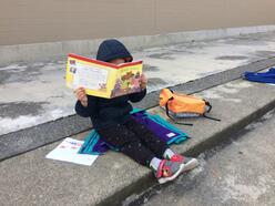 An elementary school student in a blue jacket sits on concrete steps, reading a book which covers their face. Their backpack and homework sit beside them on the concrete steps.
