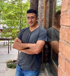 Heydar leans on a brick wall with his arms crossed in front of his chest, looking at the camera with a slight smile.
