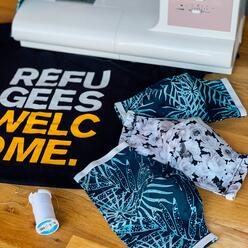 A "Refugees Welcome" Tshirt lies beneath a sewing machine and three indigo, patterned, cloth facemasks. 