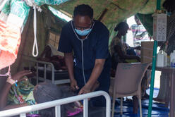 A health worker wearing PPE examines a woman stretched out on a cot at an IRC health center in Sudan.