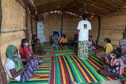 An IRC staff member wearing an IRC logo t-shirt speaks with children and adults sitting on mats on the floor in an IRC safe space for Tigrayan refugees in Sudan. 
