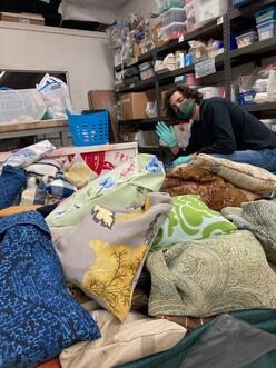 A young masked and gloved man waves and smiles at the camera, crouching andsurrounded by piles of hygiene pouches and shelving full of sewing and crafting supplies