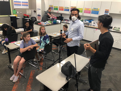 Milad and an IRC volunteer talk to seven young students in a classroom, holding video equipment with them