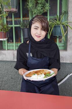 Wearing an apron, Hafeza stands in front of a building holding a plate of Afghan Bolani. 