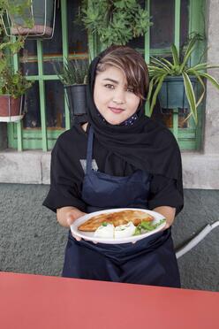 Wearing an apron, Hafeza stands in front of a building holding a plate of Afghan Bolani.