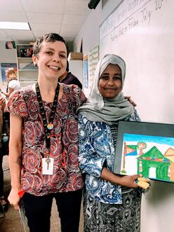 Wende stands with a young woman holding a painting of a house who is a refugee client. They are next to a whiteboard in an Atlanta classroom.