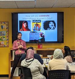 Wende stands by a projection screen with International Women's Day portraits talking to seated refugee clients.