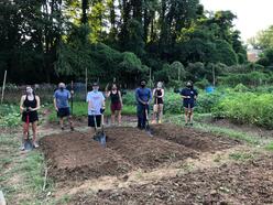 Men and women stand outside, holding garden tools, spaced out in front of three freshly dug garden beds