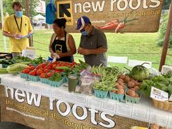 Two men and a women stand behind a table under a tent at a farmers' market display. The table is stacked with produce for sale.