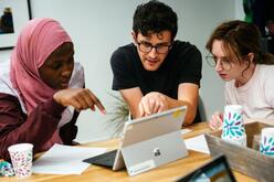 Three people hovering over a windows computer. One person pointing. Two people looking at the screen. 