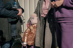 A young girl wearing a winter coat and hat leans on a woman standing next to her. 