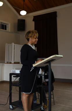 Shelleyan Lewars standing and reading from behind a podium in a large room. There is a woman sitting behind her operating a projector for an out-of-frame screen.