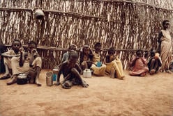 A group of young Ethiopian refugee children sit in the dirt outside a structure made of sticks and branches.