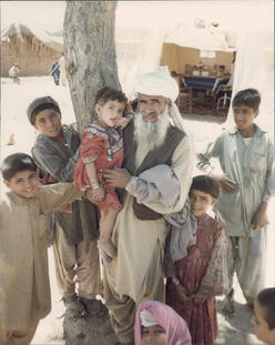 아프가니스탄 난민인 노인과 여러 어린 소년들이 나무 앞에서 사진을 찍기 위해 포즈를 취하고 있습니다.
