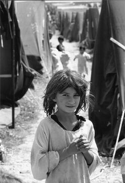 A young Iraqi refugee stands in alone, outside, in a crowded refugee camp in Turkey.