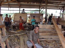 한 무리의 인도네시아인들이 큰 나무 건물에서 함께 일하며, 나무 조각들을 함께 붙여 배를 만듭니다.
