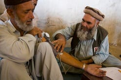 A doctor and another man, both Pakistani, sit on the floor, as the doctor takes the blood pressure of the other man.