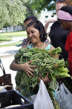 Woman holds a large bundle of greens and produce against her with a smile