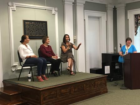 Three panelists are seated on a stage, the moderator stands at the podium to the right