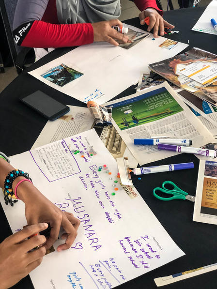 The table set with materials including magazines, scissors, markers and beads. Girls paste and decorate white papers for their dream boards.