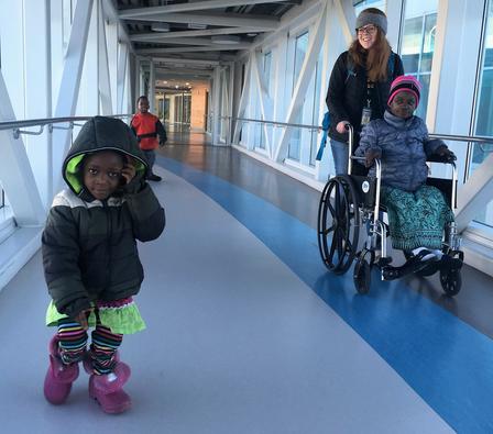 A refugee family is transported to their medical appointment by an IRC staff member An intern pushing refugee child in a wheelchair in a hospital hallway, the other two siblings walking forward.