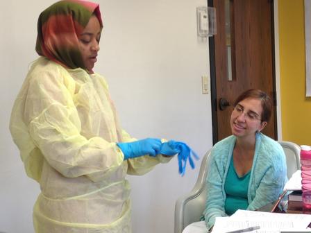 Elham passed the complete CNA test in October 2019 and is now a Certified Nursing Assistant. Student Elham puts on latex medical gloves as she stands beside her CNA instructor. She is wearing a headscarf and medical scrubs.