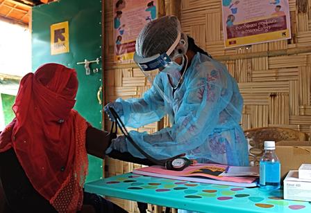 Lovely Akter, an IRC midwife, with a patient. Wearing full PPE, including a face shield, a midwife takes the blood pressure of a patient.
