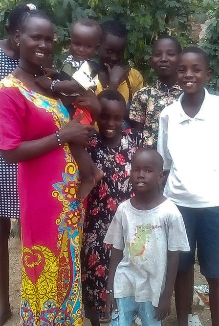Florence Lokitoe with her family. Florence Lokitoe poses with her children. They are standing outside and she is holding a baby, while five children stand next to her.