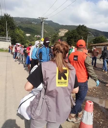 An IRC staff member stands with her back to the camera. In front of her is a line of people waiting to receive supplies. 