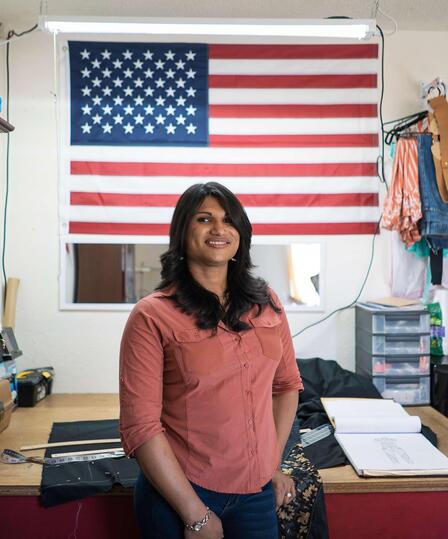 Lincy Sopall stands in her studio for her fashion design business. She is wearing a pink button down shirt and smiling. Behind her is a desk, supplies for her work, and a large American flag. 
