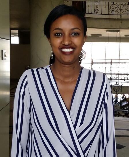 Betelhem Mengisu, wearing a black and white striped shirt and smiling at the camera, stands in a room near a spiral staircase. 