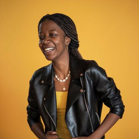 Christelle, wearing a black leather jacket, smiles and poses in front of a yellow background. 