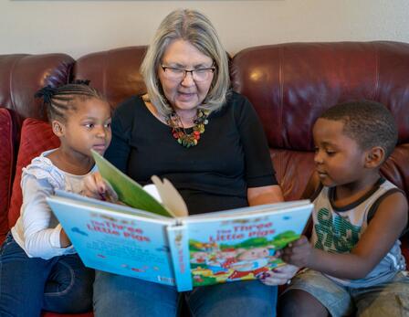 Julie Kurz reads to Sandra and Agape Julie Kurz reads to Sandra and Agape. They are sitting on a couch, the kids on either side of her with the book open on her lap.