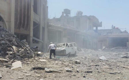 An IRC-supported hospital in the Daraa Governorate in southern Syria after it was struck by an airstrike in 2016. A man walks toward a crushed car in front of a recently bombed building. There is rubble and dust and smoke in the air.